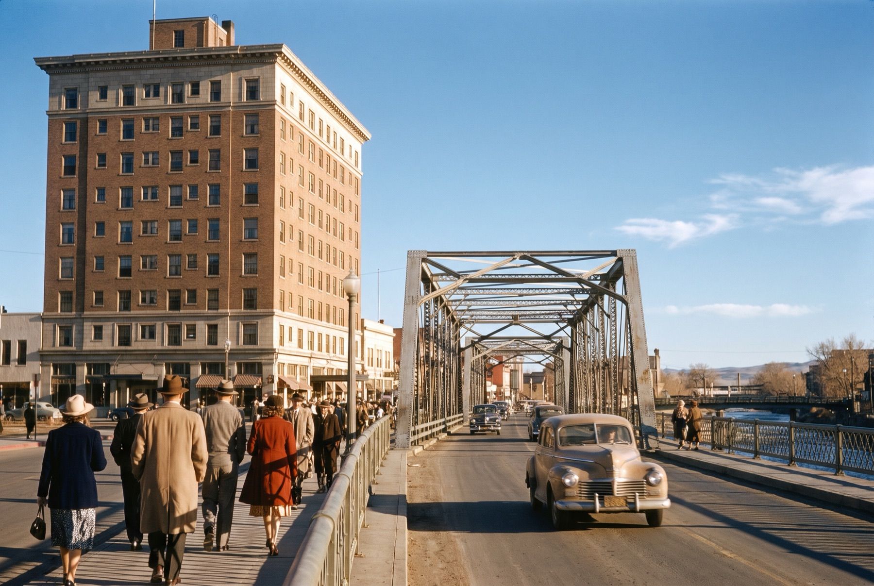 A brick hotel tower beside a steel truss bridge over the Truckee River in Reno in the late 1940s, pedestrians in overcoats on the sidewalk.