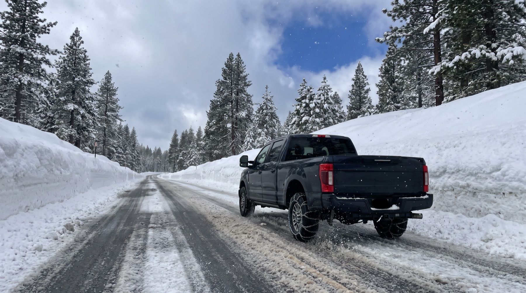 A pickup truck with heavy link chains on its rear tires climbing a snow-covered mountain highway between plowed snowbanks